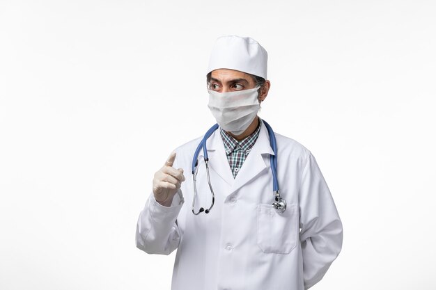 Medico maschio di vista frontale in tuta medica e maschera a causa del coronavirus sulla scrivania bianca