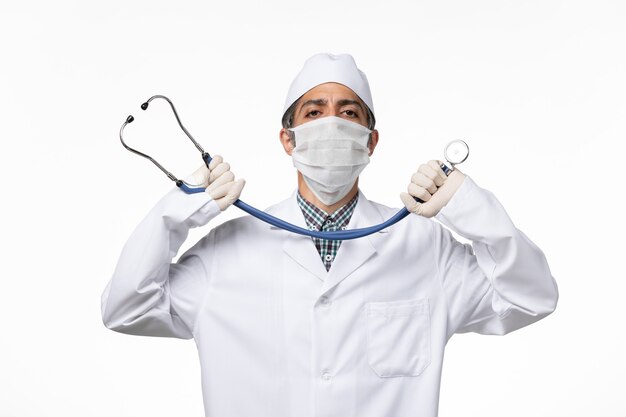 Medico maschio di vista frontale in tuta medica sterile e maschera a causa dello stetoscopio che tiene il coronavirus sulla superficie bianca