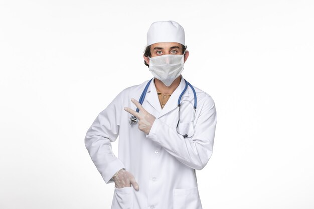 Medico maschio di vista frontale in vestito medico che indossa la sua maschera contro il coronavirus sulla medicina pandemica del virus del coronavirus della scrivania bianca