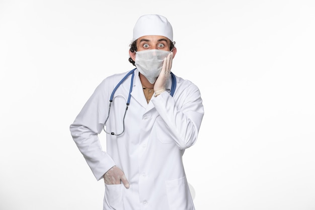Medico maschio di vista frontale in vestito medico che indossa maschera sterile come protezione da covid sulla salute della malattia pandemica del coronavirus del virus della scrivania bianca