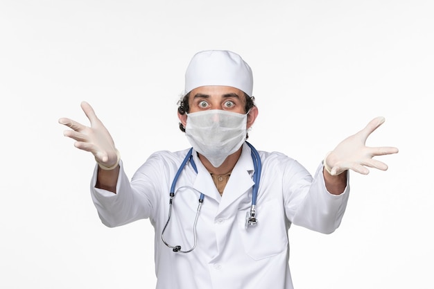 Medico maschio di vista frontale in vestito medico che indossa maschera sterile come protezione da covid sulla salute pandemica del coronavirus del virus della spruzzata della scrivania bianca