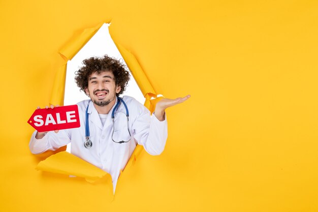 Medico maschio di vista frontale in vestito medico che tiene la scrittura di vendita sulla medicina di salute di colore del medico dello shopping giallo