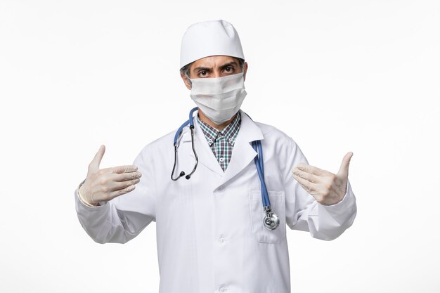 Medico maschio di vista frontale in vestito medico con maschera a causa del coronavirus sulla superficie bianca