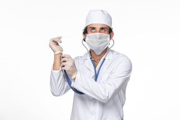 Medico maschio di vista frontale in vestito medico con maschera come protezione da covid - controllando il suo polso sulla medicina pandemica del virus del coronavirus della parete bianca