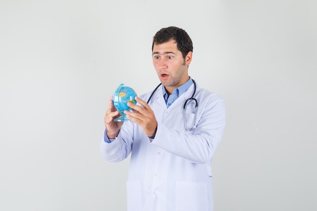 Medico maschio in camice bianco che tiene il globo del mondo e che sembra sorpreso
