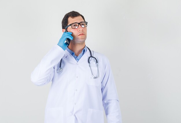 Medico maschio in camice bianco, guanti, occhiali che osserva in su mentre parla sul telefono e che sembra pensieroso