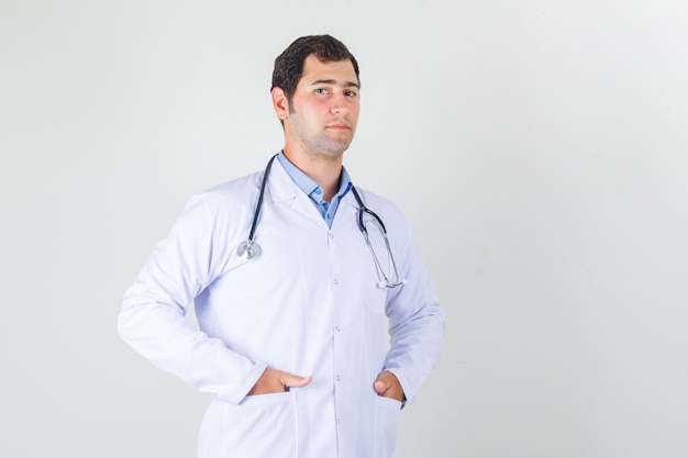 Medico maschio in piedi con le mani in tasca in camice bianco e guardando fiducioso. vista frontale.