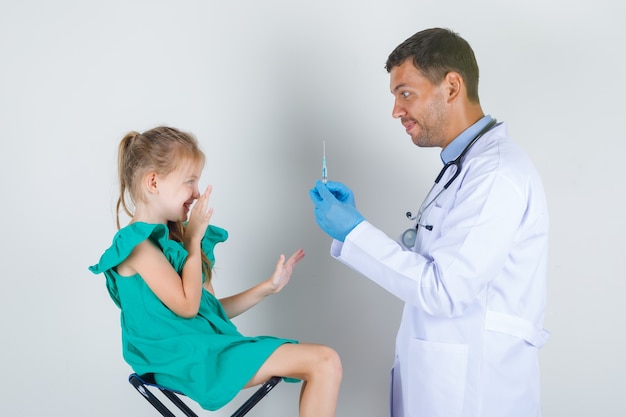 Medico maschio in uniforme bianca, guanti che tengono la siringa mentre il bambino guarda con paura