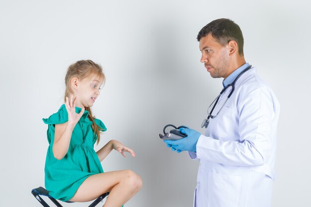 Medico maschio in uniforme bianca, guanti che tengono sfigmomanometro mentre bambino guarda con disgusto