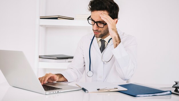 Medico maschio stanco che lavora al computer portatile in clinica