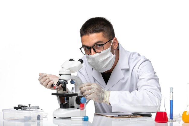 Medico maschio vista frontale in tuta medica bianca con maschera a causa di covid che lavora con il microscopio su uno spazio bianco