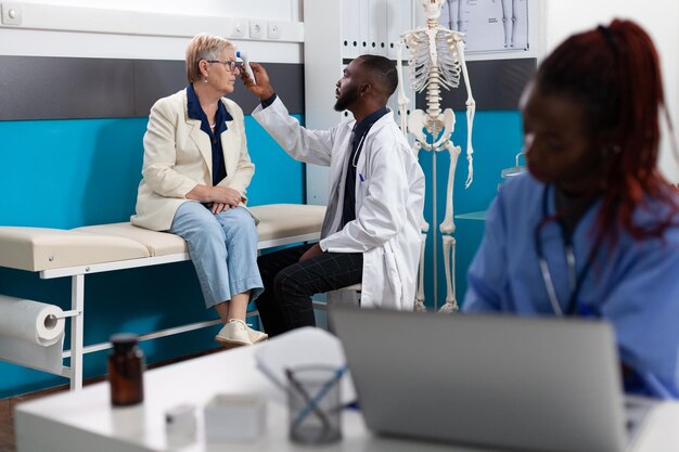 Medico medico che mette un termometro a infrarossi medico sulla fronte del paziente anziana che controlla la temperatura discutendo i sintomi della malattia durante l'appuntamento medico nell'ufficio ospedaliero. Concetto di medicina