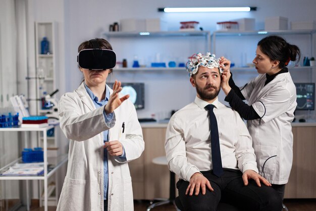 Medico medico con auricolare per realtà virtuale mentre donna neurologa che regola lo scanner eeg del paziente uomo che analizza l'evoluzione del cervello durante l'esperimento neurologico. Ingegnere scienziato che utilizza l'alta tecnologia