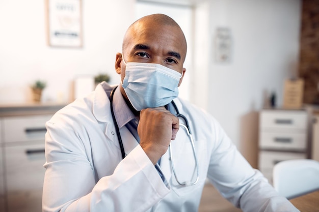 Medico nero maschio con maschera protettiva che lavora in clinica e guarda la fotocamera