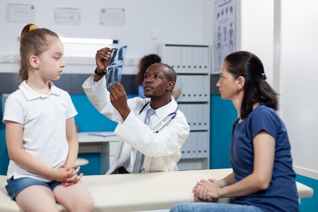 Medico pediatra afroamericano che analizza la radiografia dei polmoni discutendo i sintomi di malattia del bambino con la madre durante la consultazione medica nell'ufficio dell'ospedale. Servizio sanitario