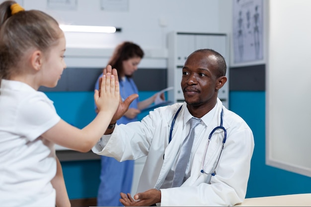 Medico pediatra afroamericano che dà il cinque ai bambini piccoli durante l'appuntamento medico nell'ufficio ospedaliero. Uomo terapista che spiega i sintomi della malattia discutendo il trattamento sanitario