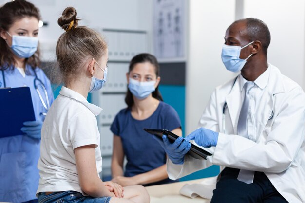 Medico pediatra afroamericano con maschera protettiva contro il coronavirus che spiega il trattamento medico alla famiglia durante l'appuntamento clinico nell'ufficio ospedaliero. Servizio sanitario