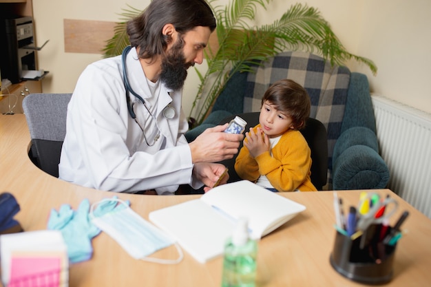 Medico pediatra che esamina un bambino in uno studio medico confortevole. Concetto di assistenza sanitaria, infanzia, medicina, protezione e prevenzione. Il ragazzino si fida del medico e si sente calmo, emozioni positive.