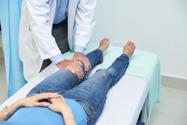 Medico potato che controlla ginocchio del paziente irriconoscibile che si trova sullo strato in ufficio medico