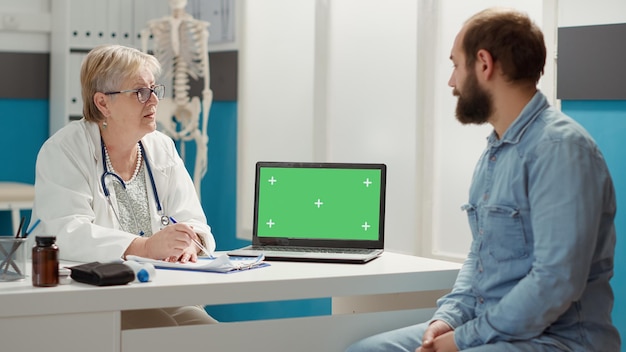 Medico senior e paziente che esaminano il laptop con schermo verde durante la visita di controllo. Uomo e donna che utilizzano un modello isolato con sfondo chromakey vuoto e spazio di copia mockup sul display.