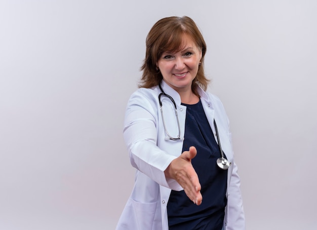 Medico sorridente della donna di mezza età che porta veste e stetoscopio medici che fanno il gesto della stretta di mano sulla parete bianca isolata con lo spazio della copia