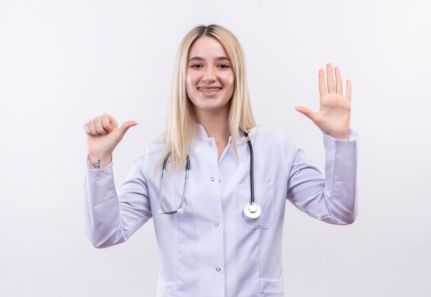 Medico sorridente giovane ragazza bionda che indossa uno stetoscopio e camice medico in parentesi graffa dentale che mostra gesto diverso su sfondo bianco isolato