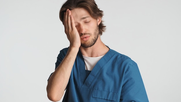 Medico stanco che tiene la mano sul viso sentendosi assonnato dopo una dura giornata in ospedale Giovane operatore medico vestito in uniforme che sembra stanco su sfondo bianco