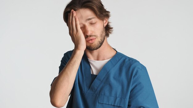 Medico stanco che tiene la mano sul viso sentendosi assonnato dopo una dura giornata in ospedale Giovane operatore medico vestito in uniforme che sembra stanco su sfondo bianco