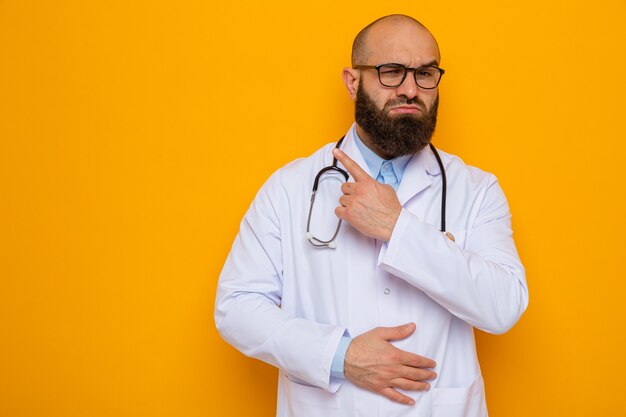 Medico uomo barbuto in camice bianco con stetoscopio intorno al collo con gli occhiali che guarda l'obbiettivo con faccia seria che punta con il dito indice al lato in piedi su sfondo arancione