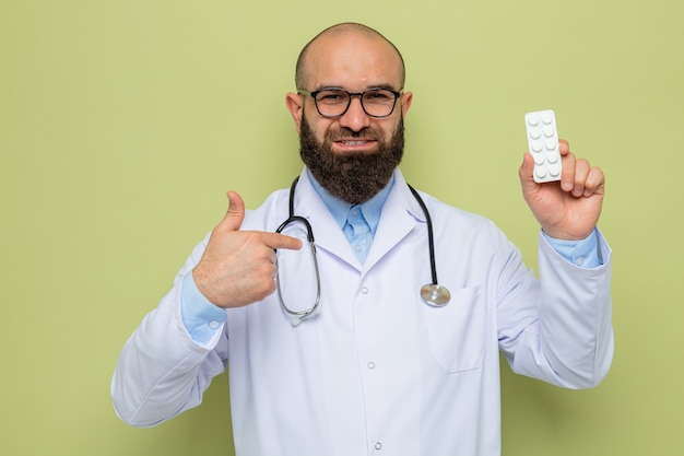 Medico uomo barbuto in camice bianco con stetoscopio intorno al collo con gli occhiali che tengono blister con pillole guardando con un sorriso sul viso felice che mostra i pollici in su