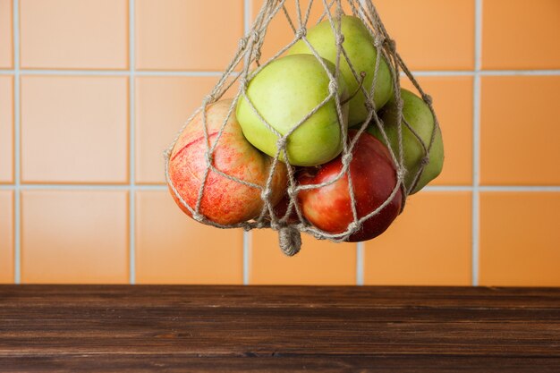 Mele che appendono in una borsa netta su un fondo di legno ed arancio delle mattonelle. vista laterale. spazio per il testo