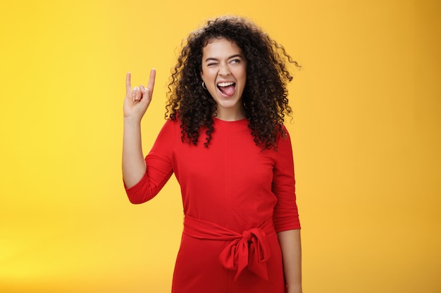 Meloman ribelle che lascia una ragazza tenera. Donna dai capelli ricci emozionata e spensierata in abito rosso che sporge la lingua e guarda bene con un sorriso mentre mostra un gesto rock-n-roll, godendo di musica fresca.