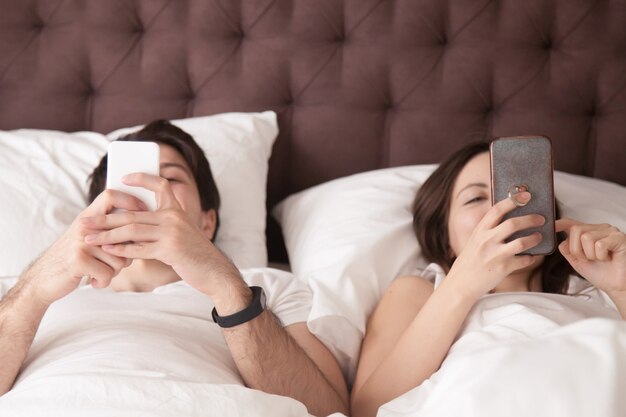 Menzogne di rilassamento delle giovani coppie a letto facendo uso degli smartphones a casa