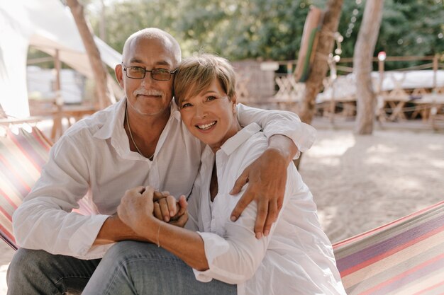 Meravigliosa donna con acconciatura bionda corta in camicetta moderna sorridente, seduto sull'amaca e abbracciando con il marito in occhiali sulla spiaggia.