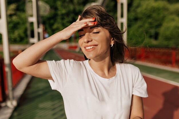 Meravigliosa donna con lunghi capelli castani che copre il viso dal sole e sorridente mentre si cammina nella giornata di sole