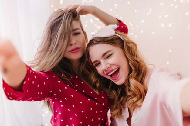 Meravigliosa donna dai capelli scuri in abito da notte rosso che si gode la mattina con un amico. Ragazza riccia estatica in maschera alla moda che fa selfie.