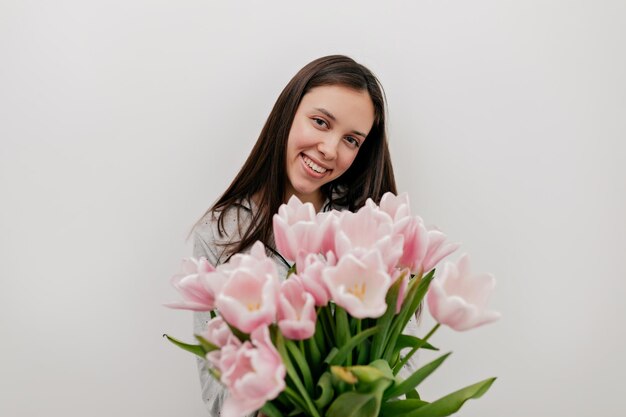 Meravigliosa donna felice con i capelli scuri e un bel sorriso felice in posa davanti alla telecamera con teneri fiori rosa su sfondo isolato La donna felice sta celebrando il giorno della donna