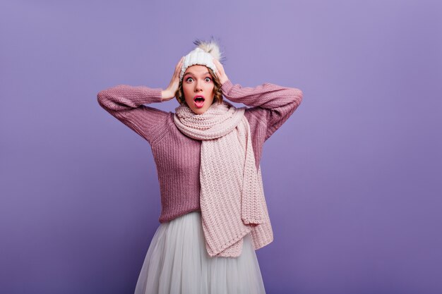 Meravigliosa donna sorpresa con sciarpa lavorata a maglia che tocca la sua testa. Ritratto dell'interno della ragazza caucasica scioccata in cappello e gonna bianca.