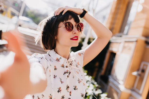 Meravigliosa ragazza bianca con labbra rosse che fa selfie mentre si cammina per la città. Splendida donna rilassata divertendosi sulla strada.