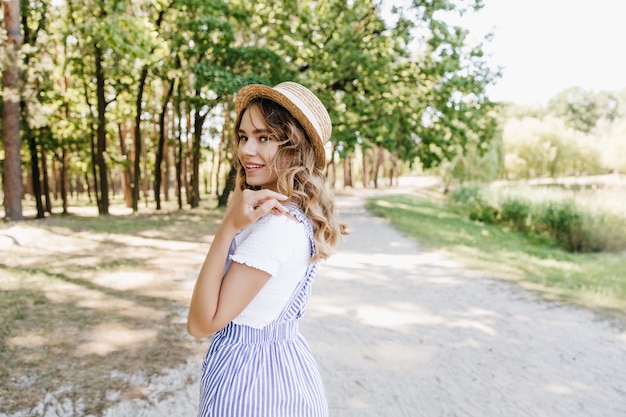 Meravigliosa ragazza bionda che cammina nel parco estivo e sorridente. Foto all'aperto di adorabile signora in cappello di paglia in posa con piacere sulla natura.