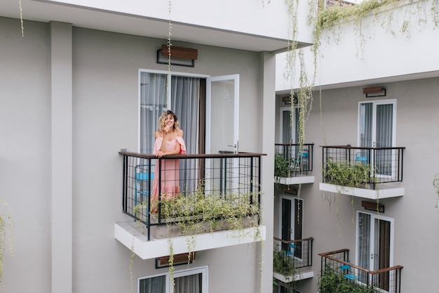 Meravigliosa ragazza caucasica che esprime felicità mentre posa in hotel. spensierata donna abbronzata riccia in abito rosa.