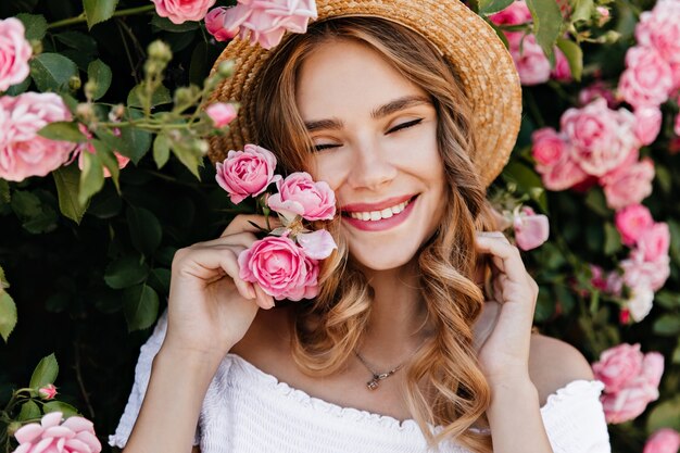 Meravigliosa ragazza caucasica in cappello di paglia che esprime felicità. Modello femminile sveglio che sta vicino al cespuglio di rose nel giorno di estate.