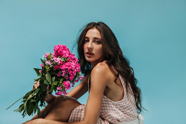 Meravigliosa ragazza con capelli ondulati in moderni abiti leggeri a quadri che tengono fiori rosa e guardano nella fotocamera su sfondo isolato