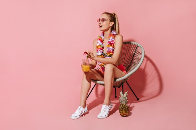 Meravigliosa ragazza con i capelli biondi in costume da bagno, occhiali da sole e collana di fiori seduta su una sedia e con in mano un cocktail sul muro rosa