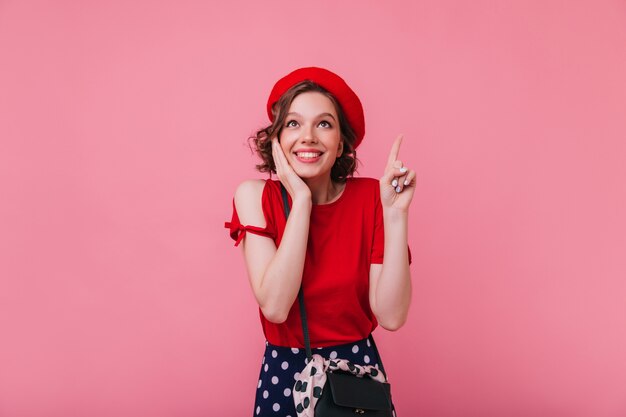 Meravigliosa ragazza francese con acconciatura ondulata in posa con un sorriso sorpreso. Foto dell'interno di graziosa donna bianca in berretto rosso isolato.