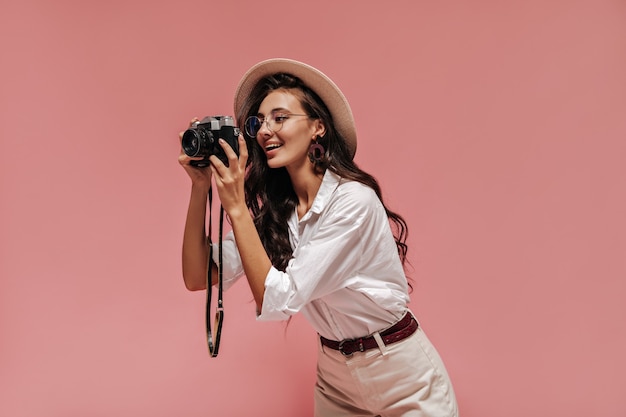 Meravigliosa signora alla moda con un'acconciatura alla moda in orecchini marroni, occhiali trasparenti e abito bianco in posa e tenendo la macchina fotografica