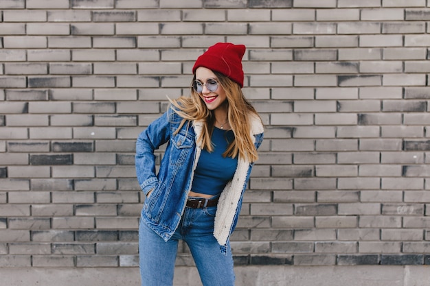 Meravigliosa signora bianca con cappello rosso che balla felice per strada. Foto all'aperto di bella ragazza bionda in vestiti di jeans divertendosi sul muro di mattoni.