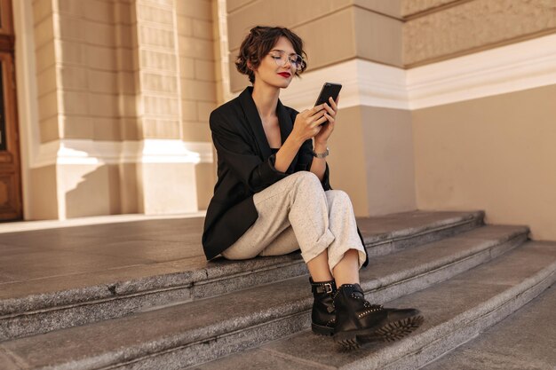 Meravigliosa signora in giacca scura e pantaloni bianchi che tiene il telefono all'esterno Donna dai capelli corti con gli occhiali seduta e sorridente all'aperto