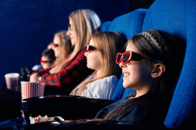 Messa a fuoco selettiva del bambino che ride con gli occhiali 3d, mangiare popcorn e guardare film divertenti. Bambina sveglia che gode del tempo con gli amici nel cinema