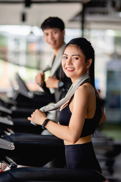 Messa a fuoco selettiva, giovane donna sexy che indossa abbigliamento sportivo e smartwatch, giovane uomo sfocato, stanno correndo sul tapis roulant per allenarsi in palestra moderna, sorriso,